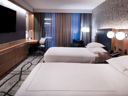 The image shows a modern hotel room with two beds, a large TV, desk, chair, lamps, and patterned wallpaper. The decor is sleek and contemporary.