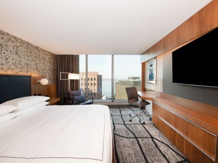 A modern hotel room with a king-size bed, a large TV, a desk, and a view of buildings and water through the window.
