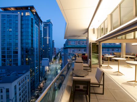 A modern rooftop patio overlooking a cityscape at dusk, with furniture and a view of tall, illuminated buildings and a street below.