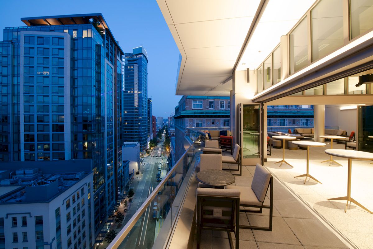 A modern balcony overlooking a city street at dusk, with tables and chairs. The cityscape features tall glass buildings and evening lights.