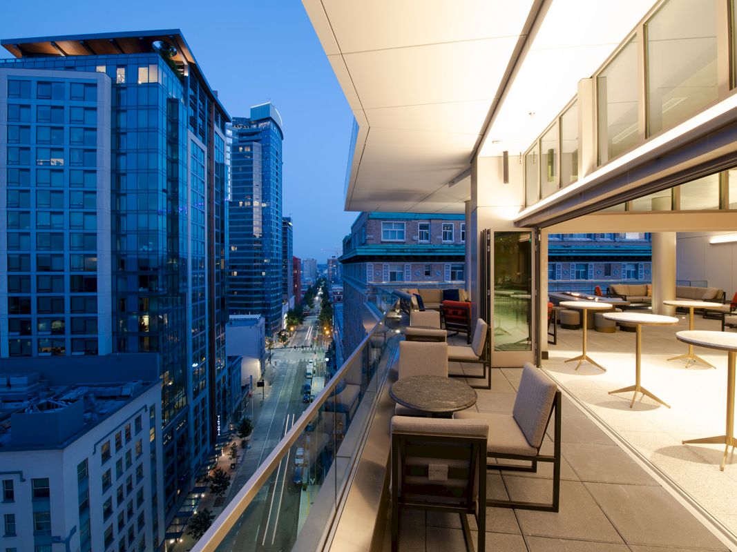 A modern balcony overlooking a city street at dusk, with tables and chairs. The cityscape features tall glass buildings and evening lights.
