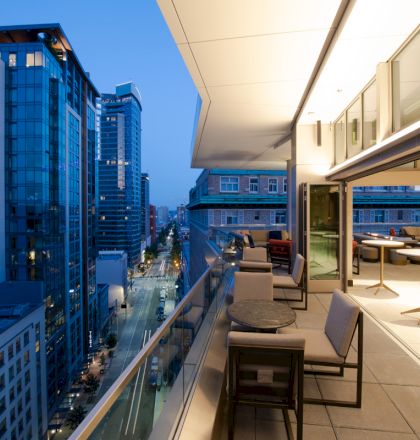 A rooftop terrace at dusk shows modern furniture and city skyscrapers. The scene captures the urban landscape with a cozy outdoor space.
