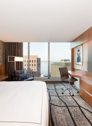 A modern hotel room with a large bed, desk, and television. The room features a window with a cityscape view and stylish decor.