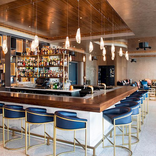 A modern bar with a central countertop, blue stools, and hanging lights; shelves are filled with bottles, and there's seating in the background.