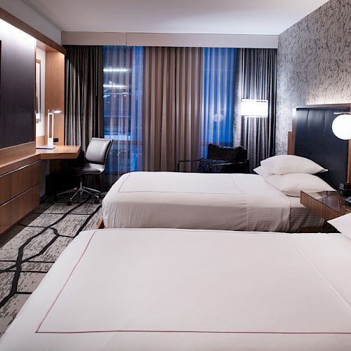 The image shows a modern hotel room with two beds, a large TV, a desk, a chair, and stylish lighting and decor.