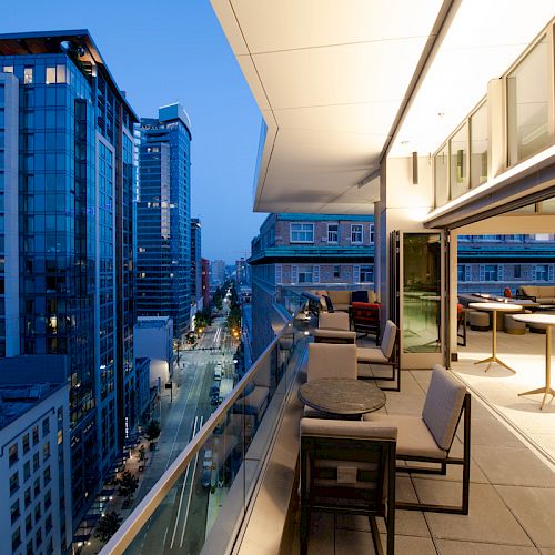 A modern cityscape at dusk with tall buildings and a stylish outdoor terrace featuring tables and chairs.