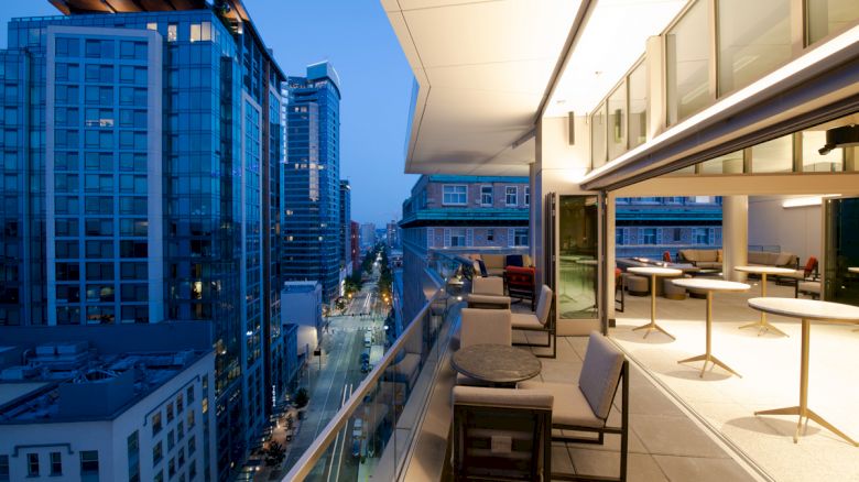A modern balcony with beige seating overlooks a cityscape featuring skyscrapers and a tranquil evening street view.