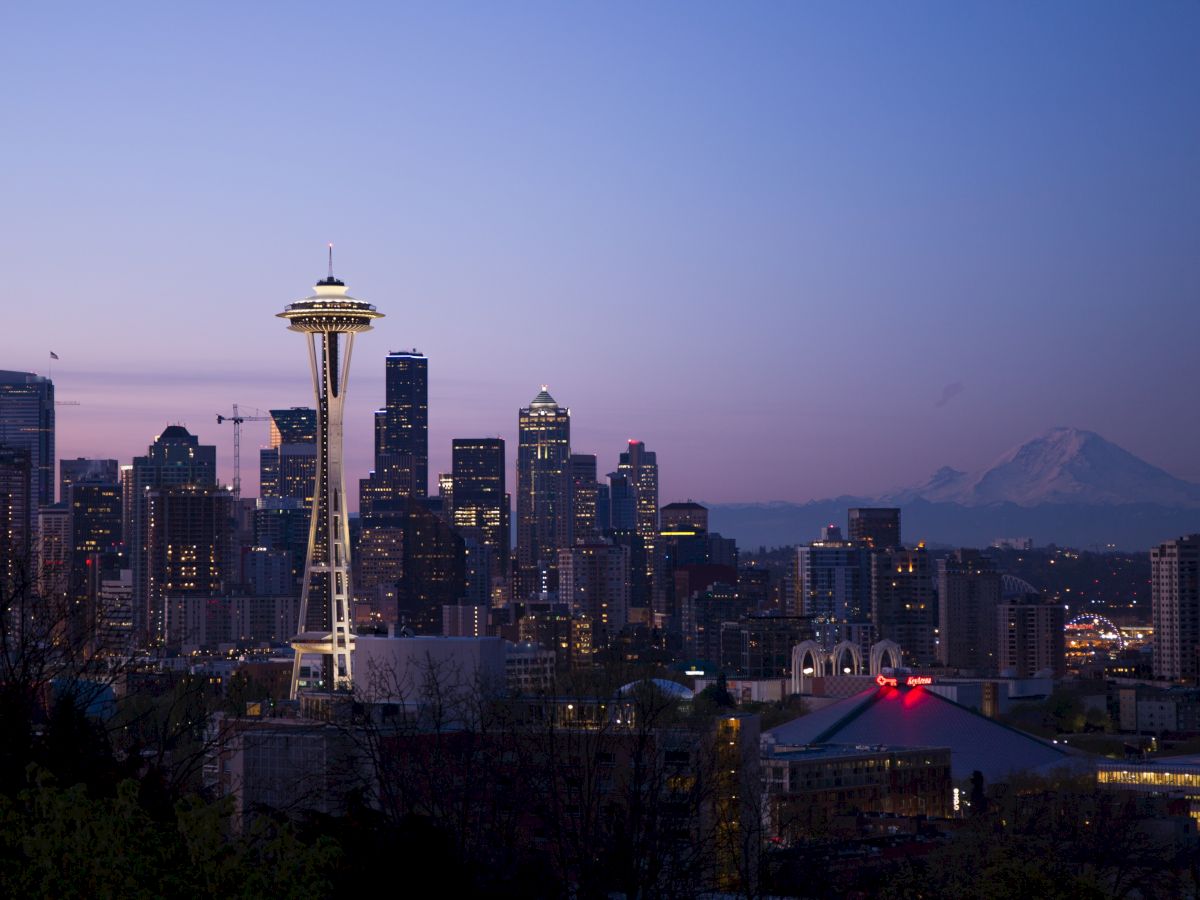 The image shows a city skyline at dusk with a prominent tower and mountains in the background, possibly Seattle with the Space Needle.