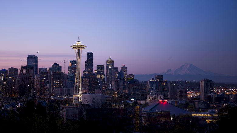 The image shows a city skyline at dusk with a prominent tower and mountains in the background, possibly Seattle with the Space Needle.
