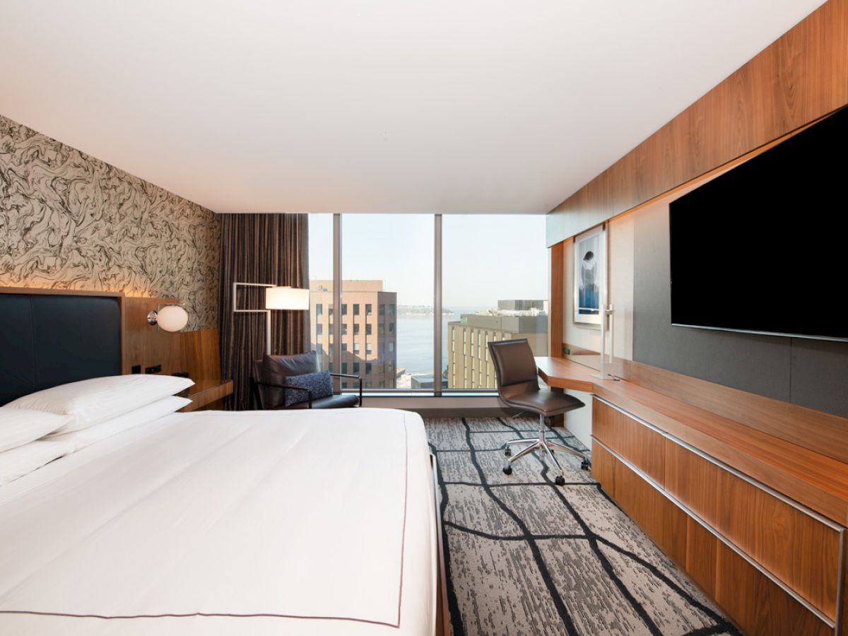 A modern hotel room with a large bed, desk, flat-screen TV, and a view of buildings through a window.