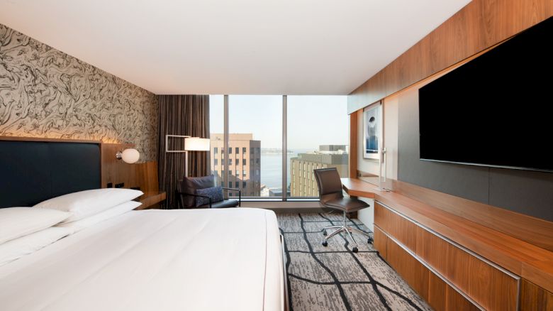 A modern hotel room with a large bed, desk, flat-screen TV, and a view of buildings through a window.