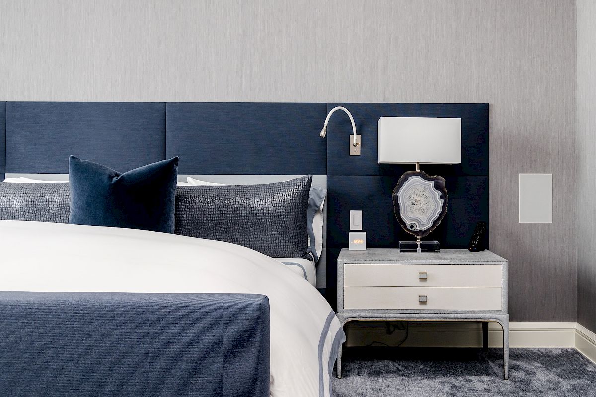 A modern bedroom features a blue headboard, white linens, a nightstand with a lamp, and a decorative item, with a neutral wall backdrop.
