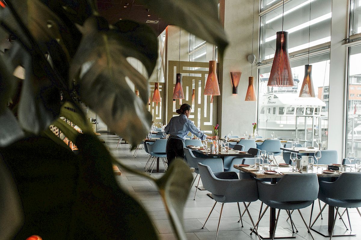 A person setting a table in a modern restaurant with stylish lamps and large windows, with leafy plants in the foreground.