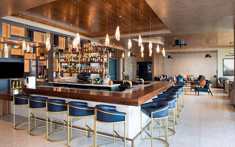 A modern bar with a wooden counter, blue stools, hanging lights, and shelves filled with bottles in a stylish, open seating area.