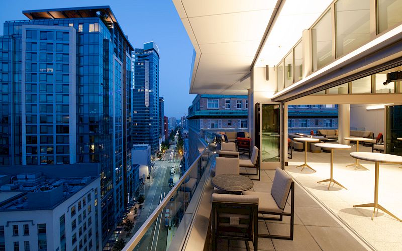 A modern cityscape with tall buildings at dusk, featuring a stylish outdoor terrace with tables, chairs, and warm lighting.