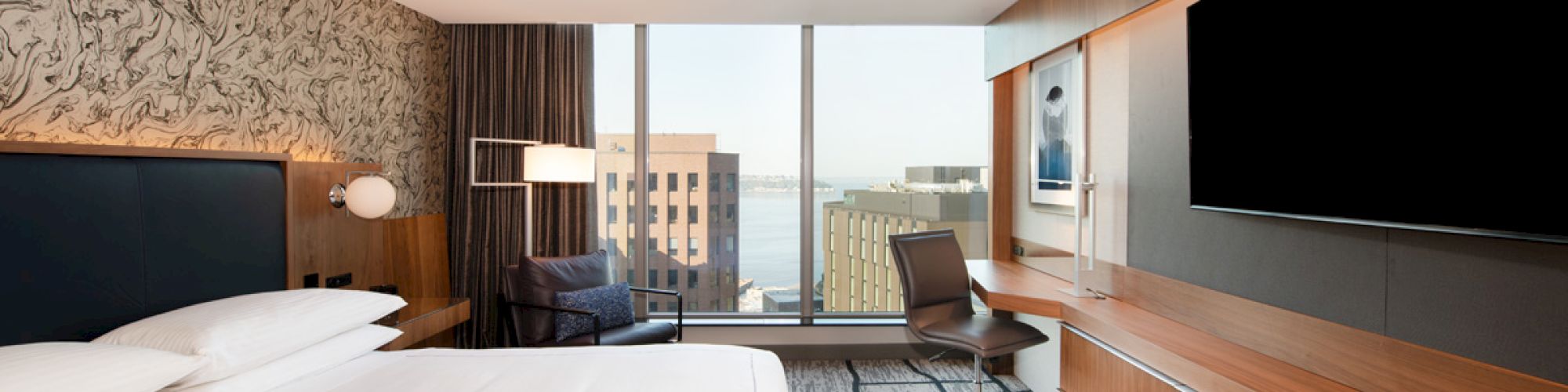 A modern hotel room with a large bed, flat-screen TV, desk, and floor-to-ceiling window offering a city view.